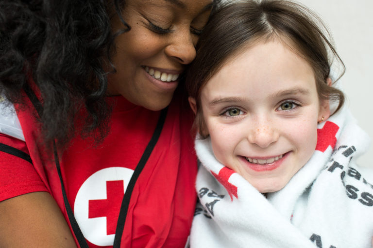 Smiling young woman embracing small girl wrapped in Red Cross blanket