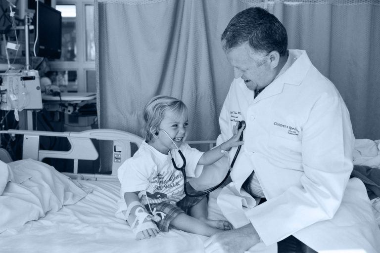 Young hospital patient uses stethoscope on older doctor on hospital bed.